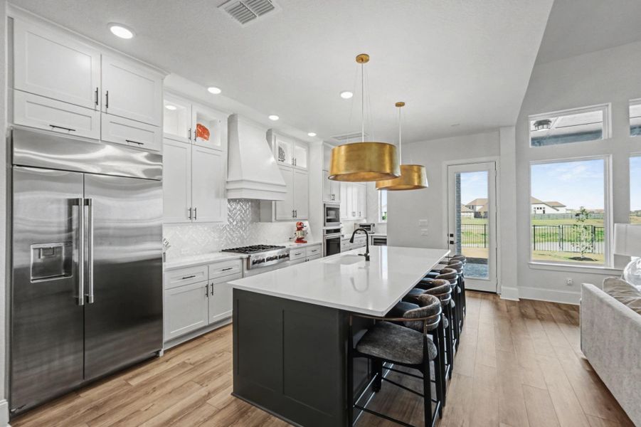 This modern kitchen features crisp white cabinets and contrasting dark cabinetry on the spacious island, all topped with elegant white quartz countertops. Integrated stainless steel appliances, including a double-door refrigerator, and a decorative tile backsplash complete the workspace. The open design overlooks the living area and outdoor patio.