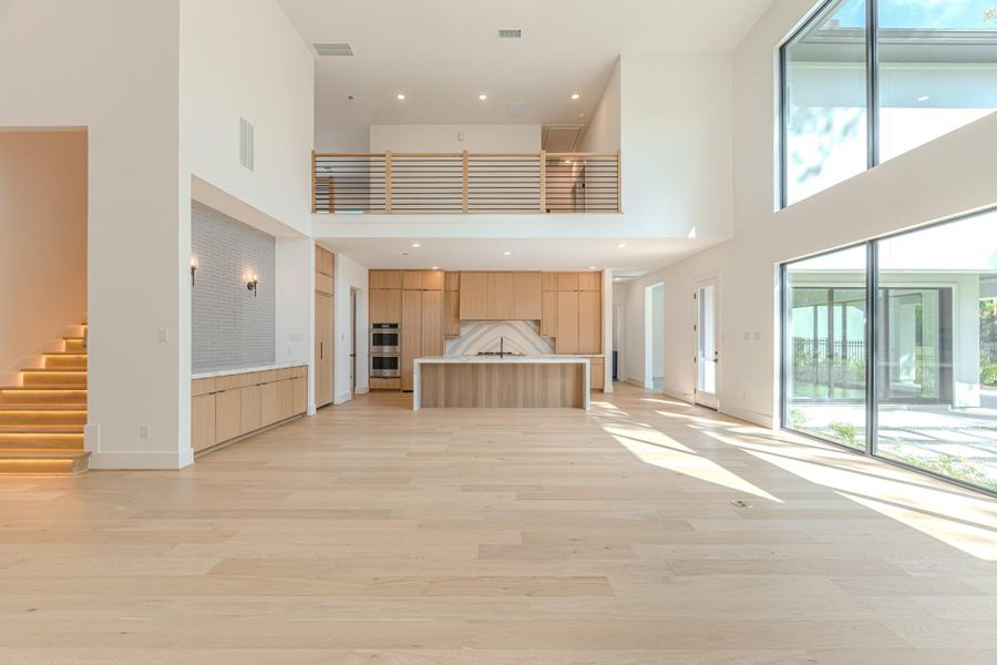 This spacious, modern living area features high ceilings, large windows for abundant natural light, and an open floor plan. The kitchen includes sleek cabinetry and a stylish island. A staircase with accent lighting leads to an upper level, enhancing the home's airy feel.