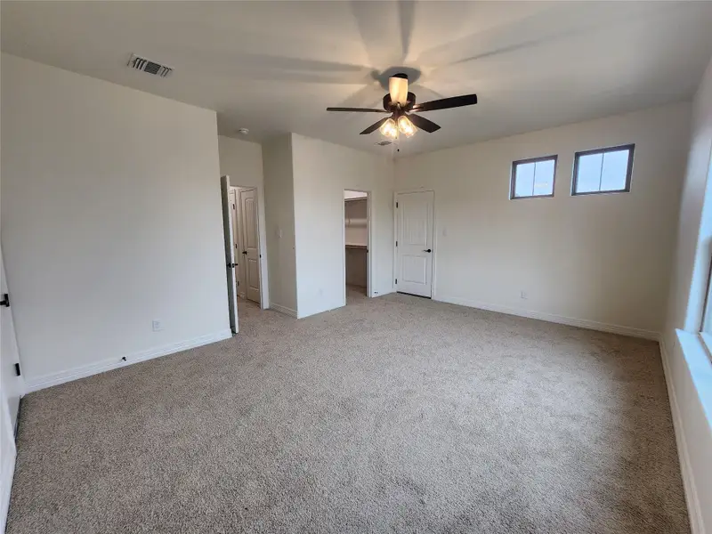 Spacious room featuring neutral carpeting, a ceiling fan with integrated lighting, two high transom windows, and white interior doors with black hardware