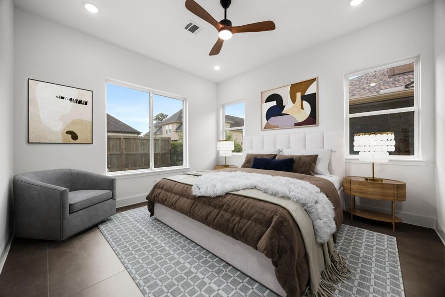 This bedroom features a modern design with a cozy bed, abstract artwork, and ample natural light from two large windows. The space includes elegant side tables, a plush gray armchair, and a ceiling fan, creating a comfortable and stylish retreat.