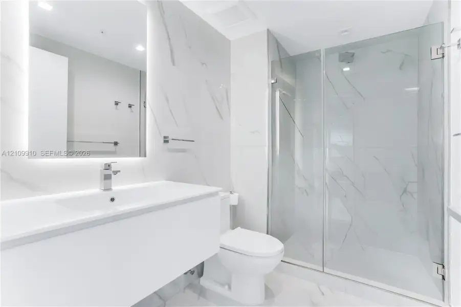 Bathroom featuring a floating vanity with a smooth surface, wall-to-wall mirror with integrated lighting, large-format stone-patterned wall tile, and a glass-enclosed shower