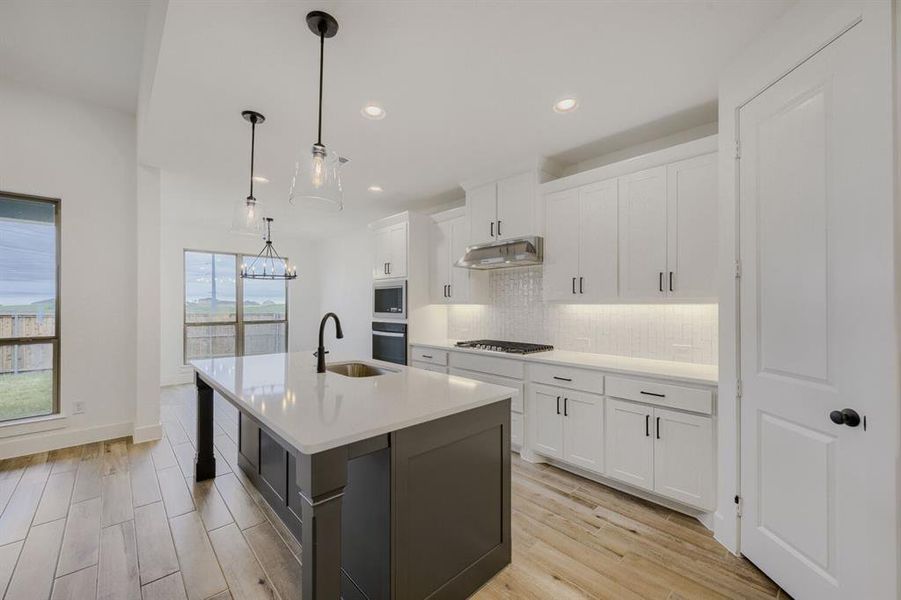 Kitchen featuring a central island with a sink and modern faucet, light-toned wood-finish flooring, white cabinetry with dark hardware, built-in stainless steel appliances, and pendant lighting