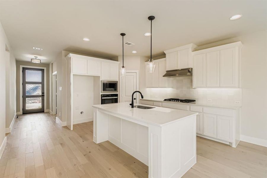 Kitchen featuring light wood finished floors, a center island with sink, white cabinetry, stainless steel appliances, and decorative light fixtures