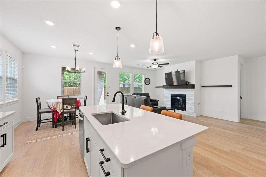 Open-concept living space with light wood-finish flooring, a kitchen island featuring a stainless steel sink and matte black faucet, recessed lighting, and a fireplace with a white tile surround and dark wood mantel Open-concept living space with light wood-finish flooring, a kitchen island featuring a stainless steel sink and matte black faucet, recessed lighting, and a fireplace with a white tile surround and dark wood mantel