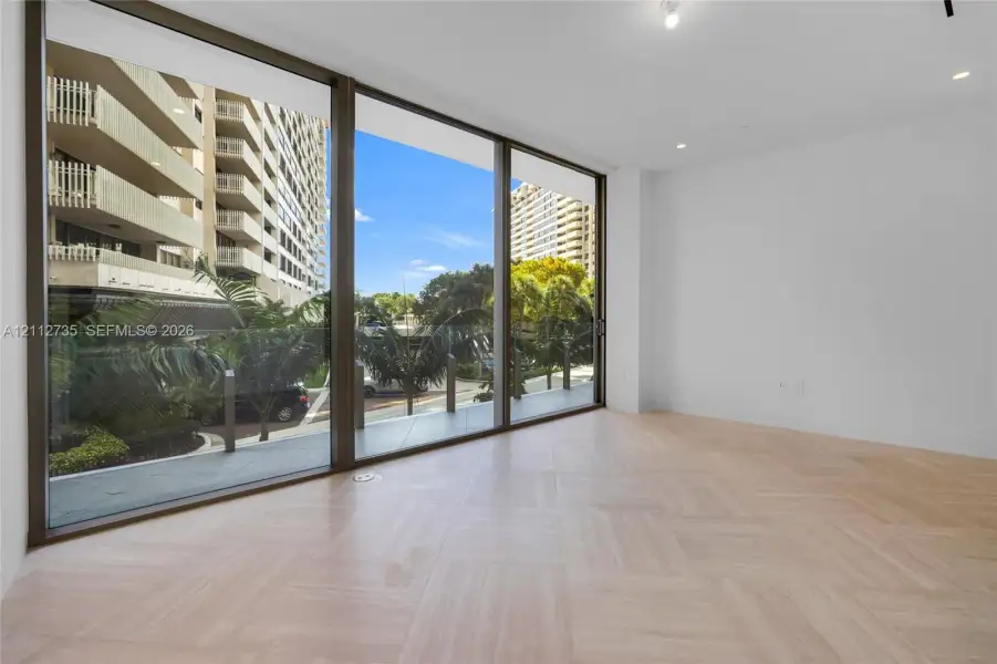 Floor-to-ceiling glass panel windows, light wood-finish herringbone flooring, recessed ceiling lighting, and a glass-railed balcony