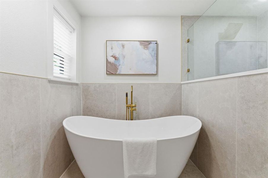 The bathroom features a freestanding bathtub with a gold-toned faucet, light-colored tiled walls, and a window with blinds The bathroom features a freestanding bathtub with a gold-toned faucet, light-colored tiled walls, and a window with blinds