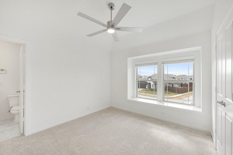 Empty bedroom with carpet flooring, ceiling fan, large window, and attached bathroom visible through open door
