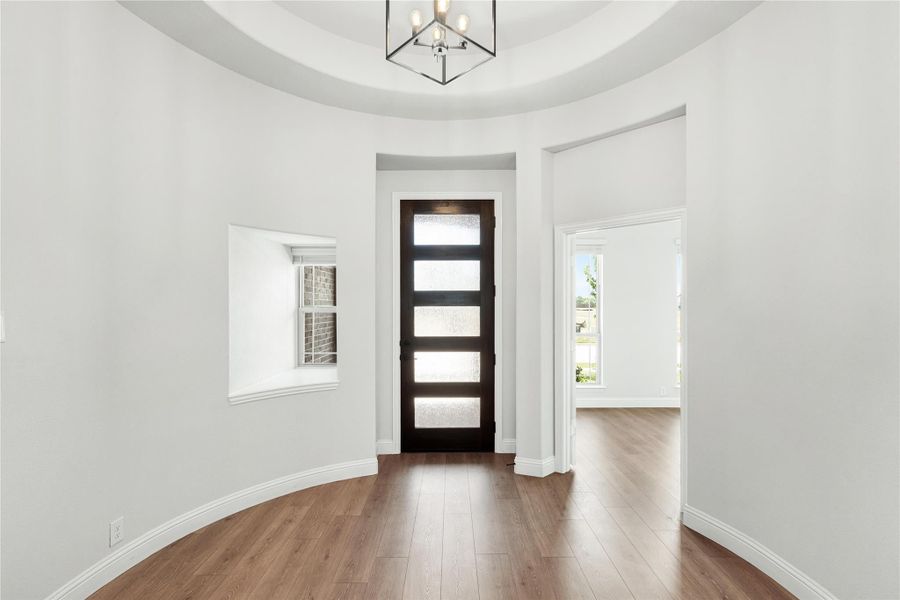 Round foyer with tray ceiling, modern chandelier, dark front door with glass panels, and hardwood floors
