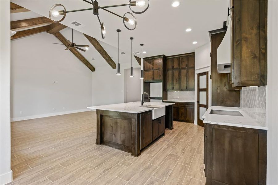 Open-concept kitchen and living area featuring vaulted ceilings with exposed wood beams, a contemporary chandelier, recessed lighting, a kitchen island with an undermount farmhouse sink, and a cooktop with a range hood