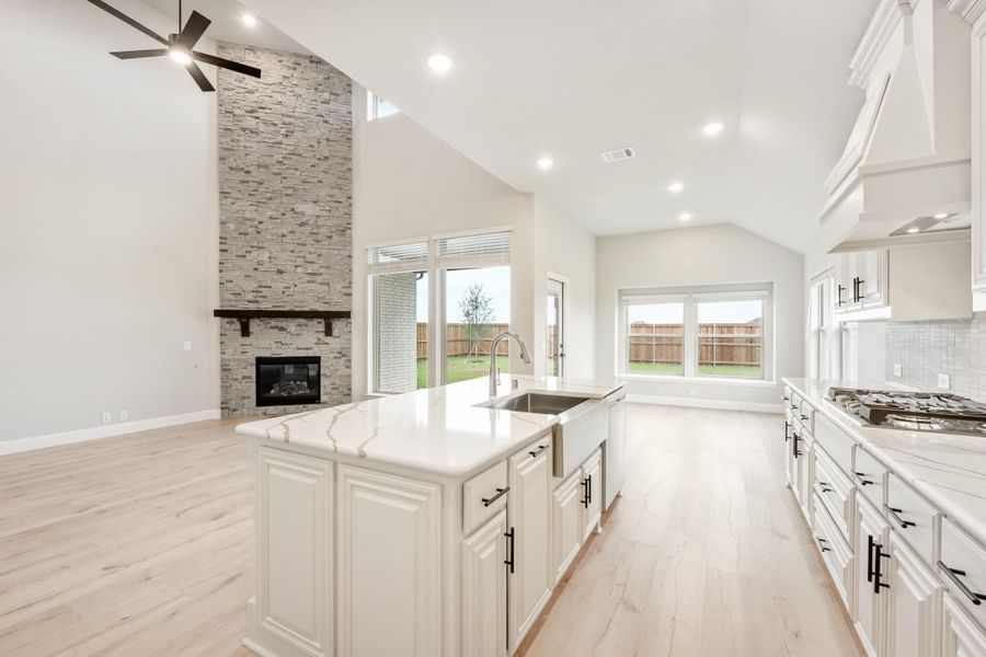 Open kitchen with white cabinets, quartz island, and stone fireplace under vaulted ceiling with light wood floors