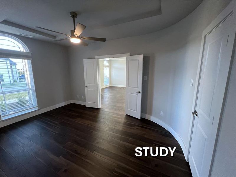 Spacious room featuring dark wood-finish flooring, a contemporary ceiling fan with integrated lighting, tray ceiling, and an arched transom window Spacious room featuring dark wood-finish flooring, a contemporary ceiling fan with integrated lighting, tray ceiling, and an arched transom window