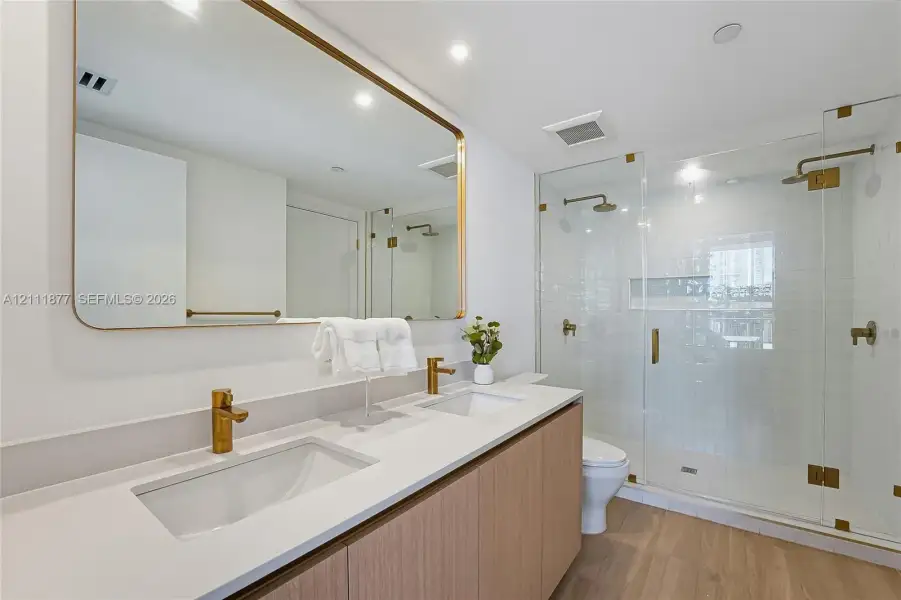 Double vanity with rectangular undermount sinks, quartz countertop, brass-finish fixtures, wood-finish cabinetry, and glass-enclosed shower with subway tile surround