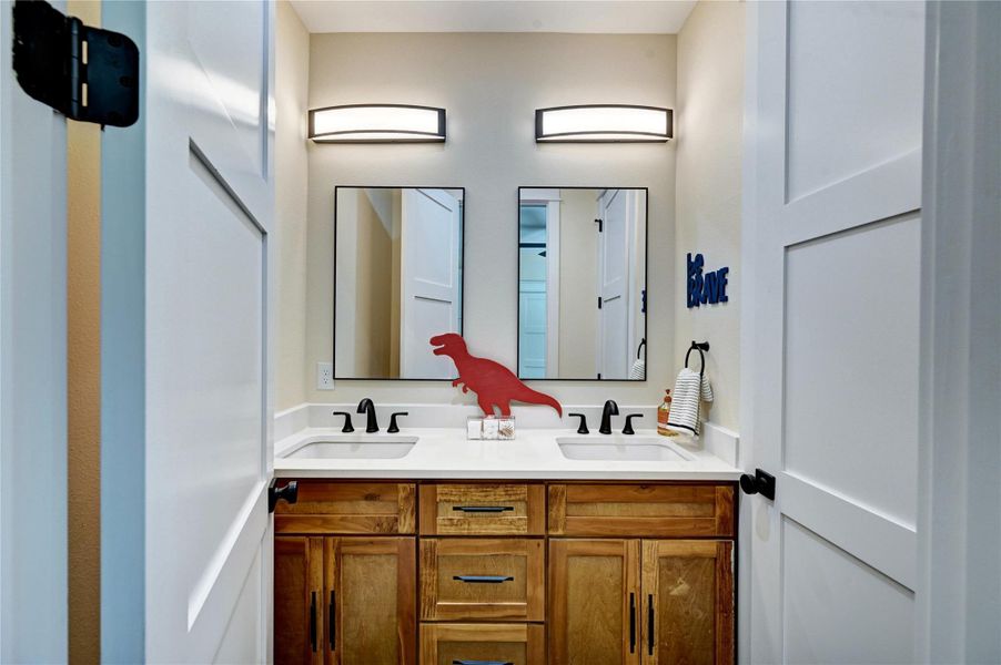 This photo showcases a modern bathroom with a double vanity featuring sleek black fixtures and wooden cabinetry. The design includes bright lighting, two mirrors, and a playful red dinosaur accessory.
