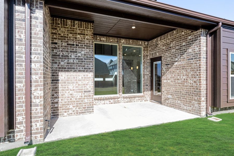 Covered back patio with brick exterior, concrete slab, large window, and back door overlooking green lawn Covered back patio with brick exterior, concrete slab, large window, and back door overlooking green lawn