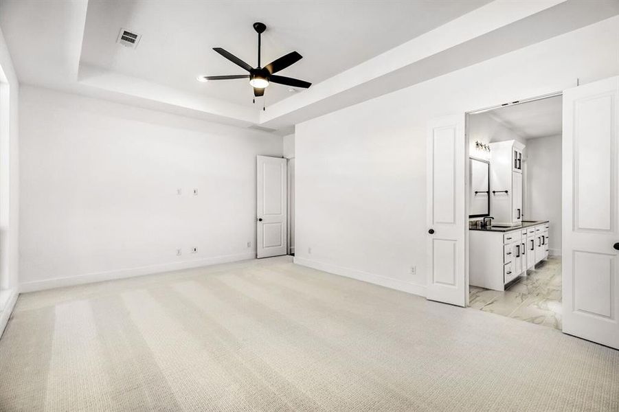 Unfurnished bedroom with light carpet, ensuite bath, ceiling fan, and a tray ceiling