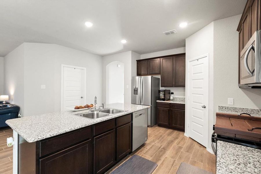 Kitchen with dark brown cabinets, stainless steel appliances, a kitchen island with sink, sink, and light hardwood / wood-style flooring