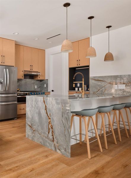 Kitchen featuring backsplash, light brown cabinetry, a peninsula, hanging light fixtures, and stainless steel fridge with ice dispenser Kitchen featuring backsplash, light brown cabinetry, a peninsula, hanging light fixtures, and stainless steel fridge with ice dispenser