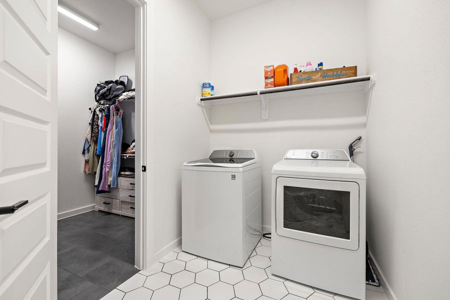 Laundry area featuring white hexagon tile flooring, a white wall-mounted shelf, and a doorway opening to a closet with dark gray tile flooring