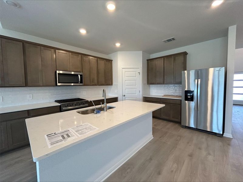 Kitchen with appliances with stainless steel finishes, dark brown cabinets, tasteful backsplash, light stone countertops, and light wood-style flooring