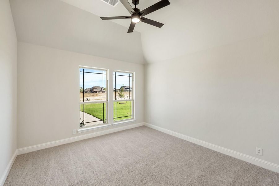 Empty bedroom with vaulted ceiling, ceiling fan, carpet flooring, and two windows overlooking a backyard