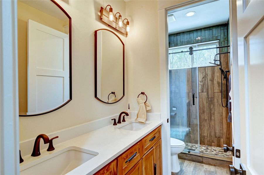 This bathroom features a double vanity with elegant fixtures, wood cabinetry, and a large mirror. The shower is enclosed with glass doors and has rustic wood-style tiles, creating a warm, inviting atmosphere.