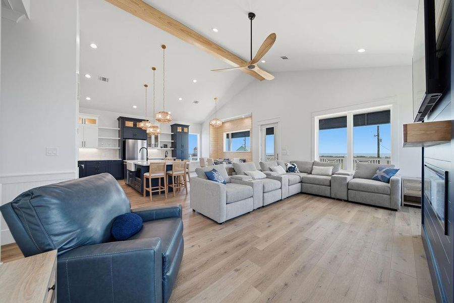 This spacious open-concept living area features high ceilings, modern lighting, and ample natural light. The kitchen boasts sleek cabinetry, a large island with seating, and stainless steel appliances. Comfortable seating and a stylish ceiling fan enhance the cozy yet contemporary vibe, while large windows offer scenic views.