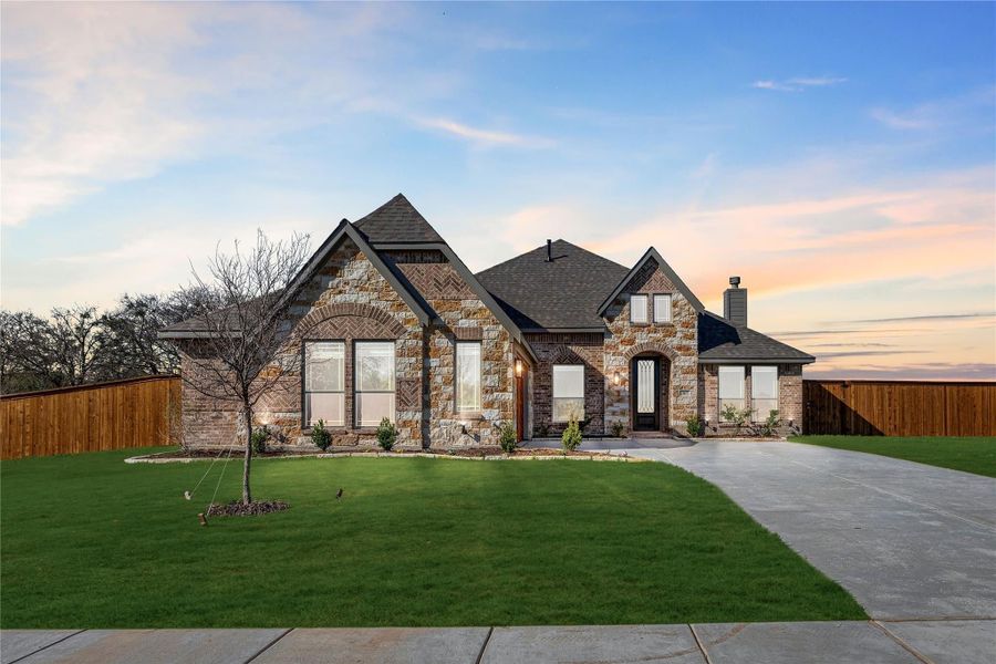 Single-story home with stone and brick exterior, arched entry, dark roof, and curved driveway with green lawn at dusk. Single-story home with stone and brick exterior, arched entry, dark roof, and curved driveway with green lawn at dusk.
