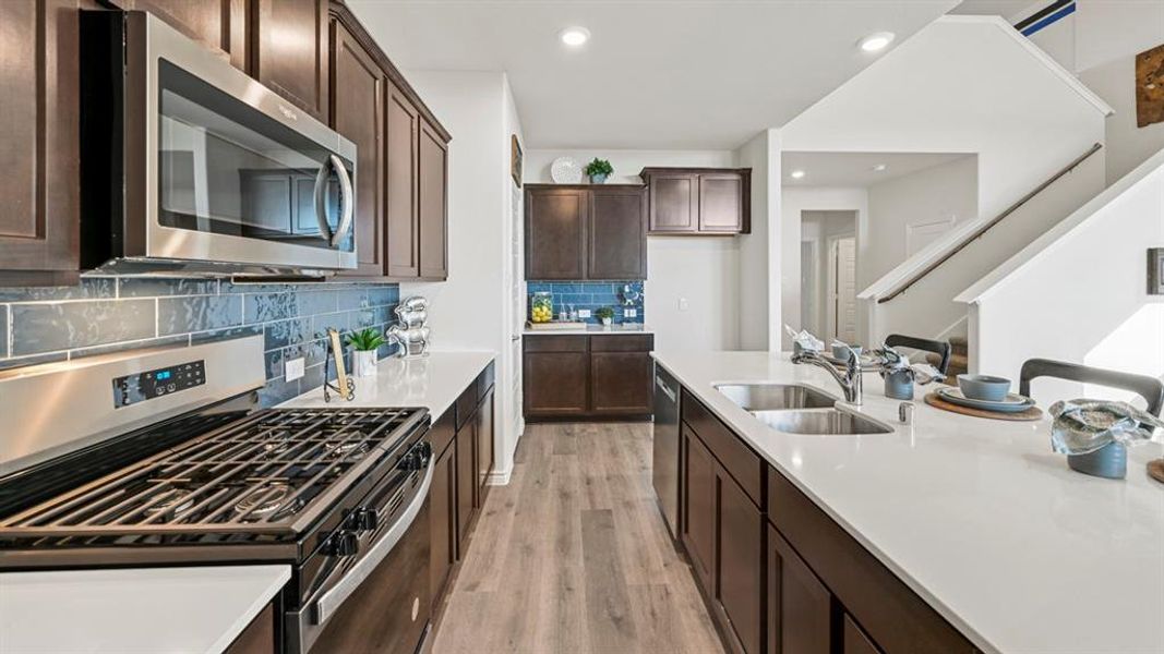 Modern kitchen featuring dark wood cabinetry, stainless steel appliances, light countertops, a subway tile backsplash, and wood-finish flooring Modern kitchen featuring dark wood cabinetry, stainless steel appliances, light countertops, a subway tile backsplash, and wood-finish flooring