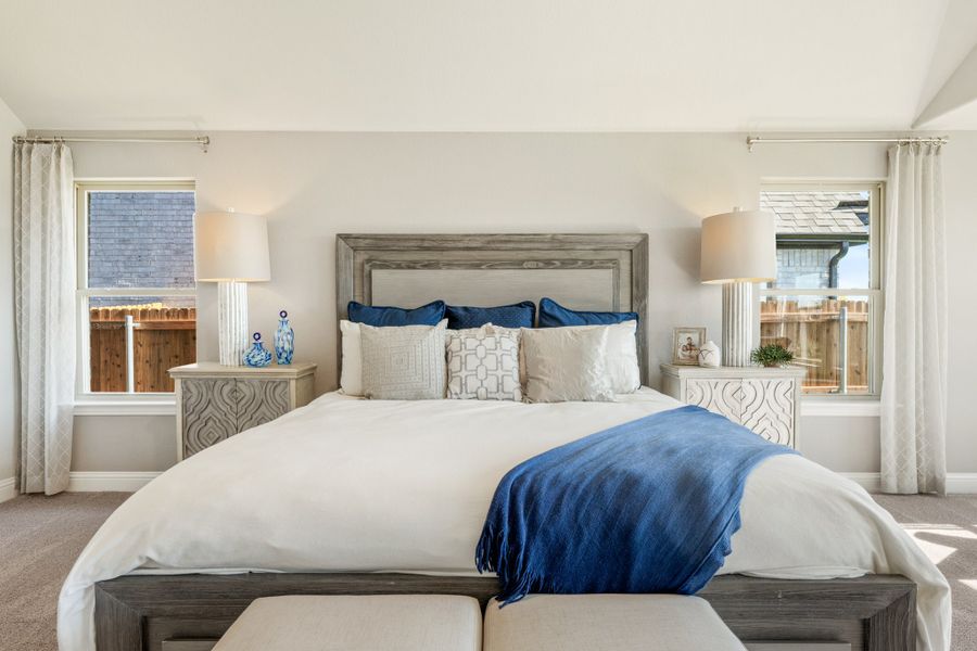 Master bedroom with gray wood headboard, white bedding, blue accents, two windows, and matching nightstands with lamps