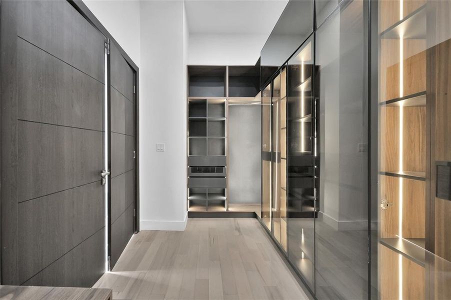 This photo showcases a sleek, modern walk-in closet with dark wood cabinetry, glass-fronted storage, and integrated lighting. The space features light hardwood flooring and a minimalist design, ideal for organized and stylish storage.