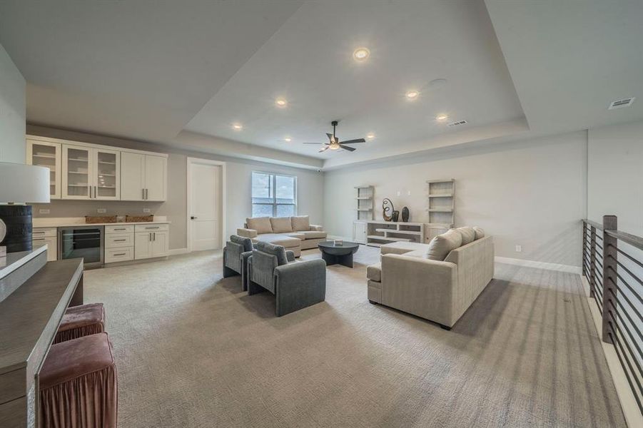 Living area with a raised ceiling, light colored carpet, beverage cooler, ceiling fan, and a dry bar Living area with a raised ceiling, light colored carpet, beverage cooler, ceiling fan, and a dry bar