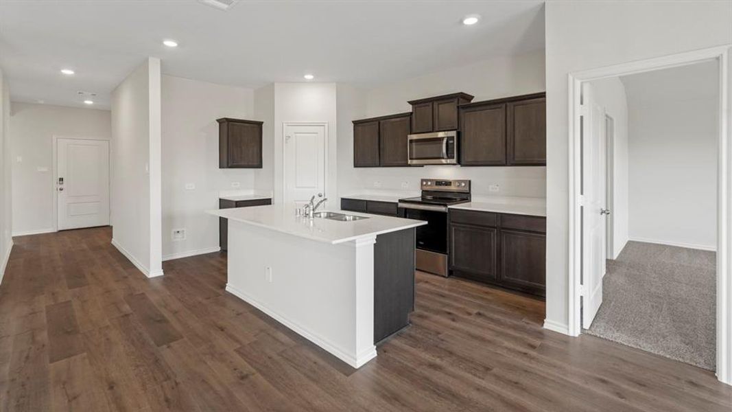 Open-concept kitchen featuring an island with an integrated sink, dark wood-finish cabinetry, stainless steel appliances, and wood-finish flooring