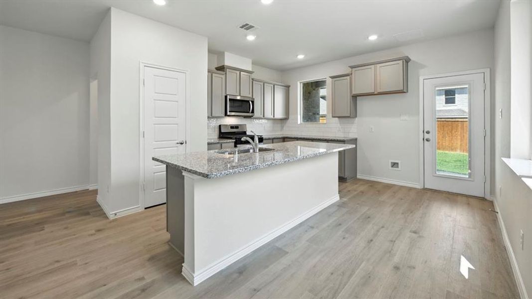 Kitchen featuring gray cabinetry, light stone counters, a center island with sink, stainless steel appliances, and backsplash