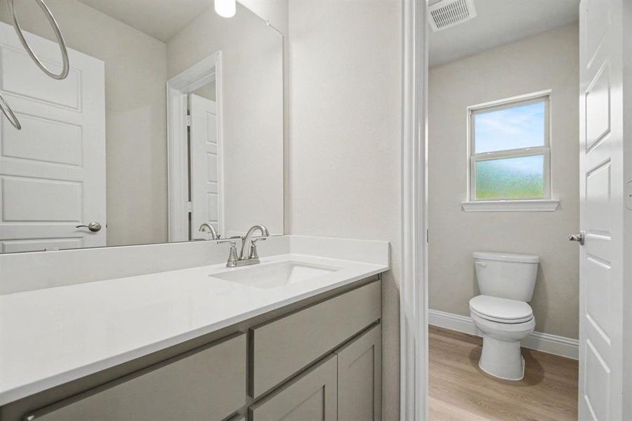 Bathroom featuring a light gray vanity with a white countertop and undermount sink, a large wall-mounted mirror, brushed nickel faucet, and wood-finish flooring Bathroom featuring a light gray vanity with a white countertop and undermount sink, a large wall-mounted mirror, brushed nickel faucet, and wood-finish flooring