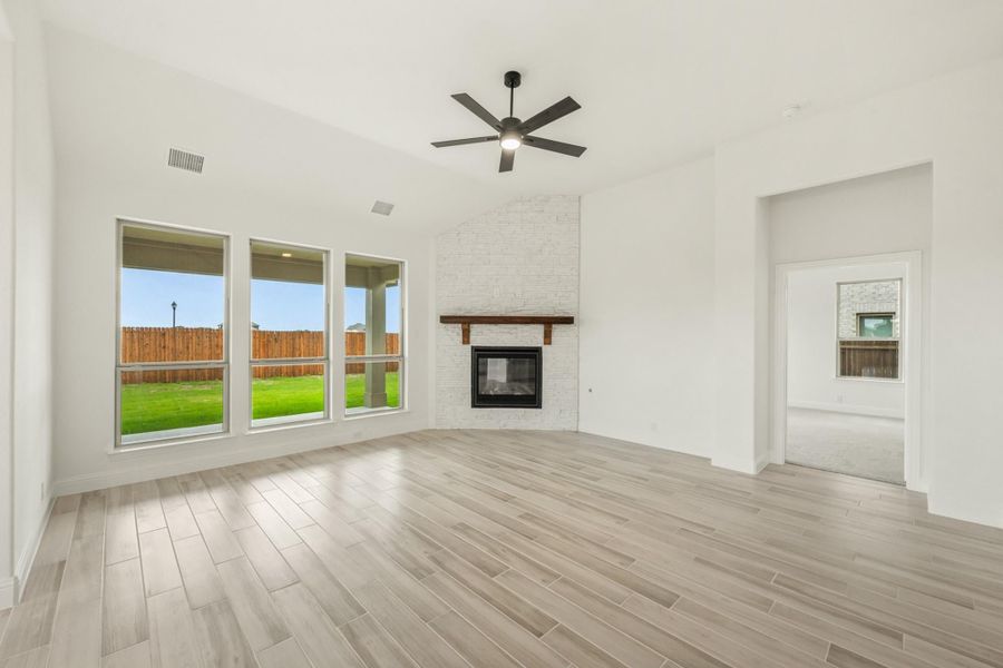 Open living room with light wood floors, brick fireplace, ceiling fan, and large windows overlooking backyard Open living room with light wood floors, brick fireplace, ceiling fan, and large windows overlooking backyard