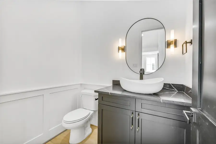 A statement of refined simplicity, this second-floor powder room features a custom floating vanity with shaker cabinetry, topped with a marble-inspired vessel sink and paired with a sleek brushed brass faucet. Elegant modern sconces flank the arched mirror, casting a soft ambient glow that complements the crisp white millwork wainscoting.Every design detail—from the hardware to the lighting—reflects thoughtful craftsmanship, seamlessly blending functionality with boutique hotel–level sophistication. A statement of refined simplicity, this second-floor powder room features a custom floating vanity with shaker cabinetry, topped with a marble-inspired vessel sink and paired with a sleek brushed brass faucet. Elegant modern sconces flank the arched mirror, casting a soft ambient glow that complements the crisp white millwork wainscoting.Every design detail—from the hardware to the lighting—reflects thoughtful craftsmanship, seamlessly blending functionality with boutique hotel–level sophistication.