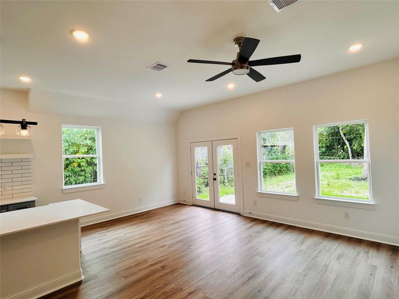 This bright and inviting living area offers the perfect blend of modern comfort and open-concept luxury. Featuring soaring ceilings, rich wood-style flooring, recessed lighting, and an abundance of natural light, the space feels both spacious and welcoming. Large picture windows and elegant French doors create a seamless indoor-outdoor flow while showcasing peaceful backyard views.