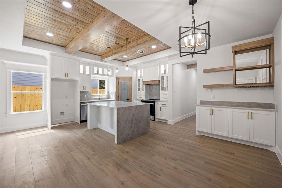 Open-concept kitchen and dining area featuring a wood-finish coffered ceiling, light wood-finish flooring, white cabinetry with gold-finish hardware, a center island with a waterfall-edge countertop, and a built-in buffet with floating wood shelves