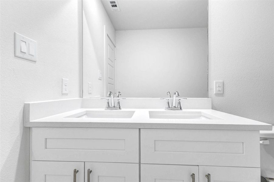 Close-up view of the secondary bath vanity with dual undermount sinks, white cabinetry, chrome fixtures, and a wide mirror. The bright neutral design provides a clean and functional space with plenty of counter area for shared daily routines.