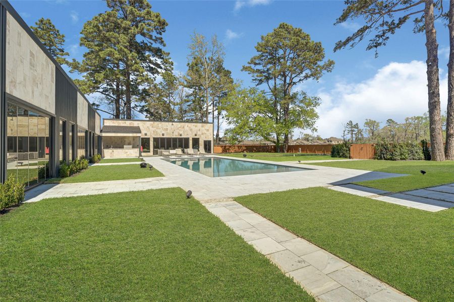 Modern backyard featuring a spacious pool surrounded by a large patio and lush green lawn. Tall trees and a wooden fence provide privacy and a natural backdrop, perfect for relaxation and outdoor entertainment.