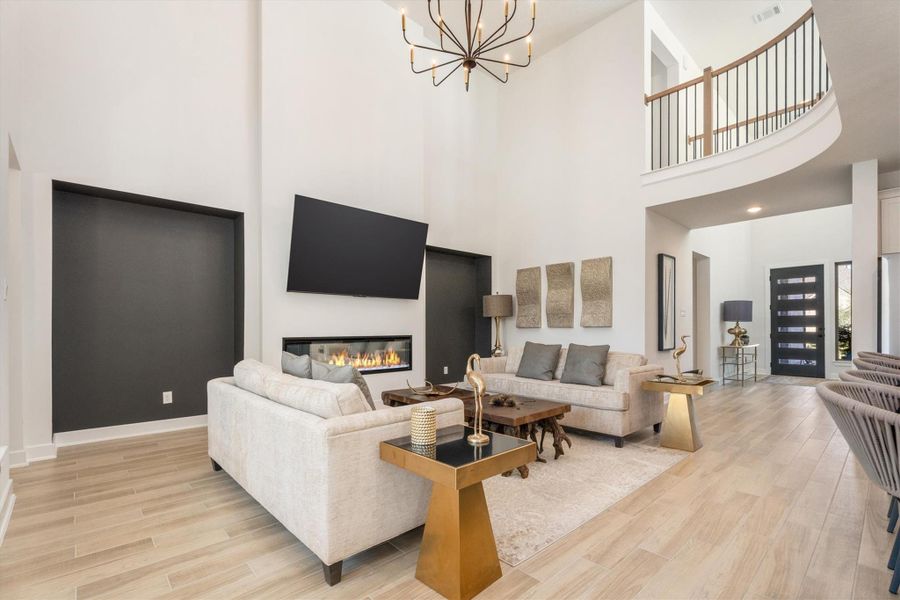This spacious living room features high ceilings, a modern chandelier, and a cozy fireplace. It includes stylish, neutral-toned furniture and a wall-mounted TV, offering a sleek, contemporary feel. The open layout seamlessly connects to the entryway with elegant wood flooring.