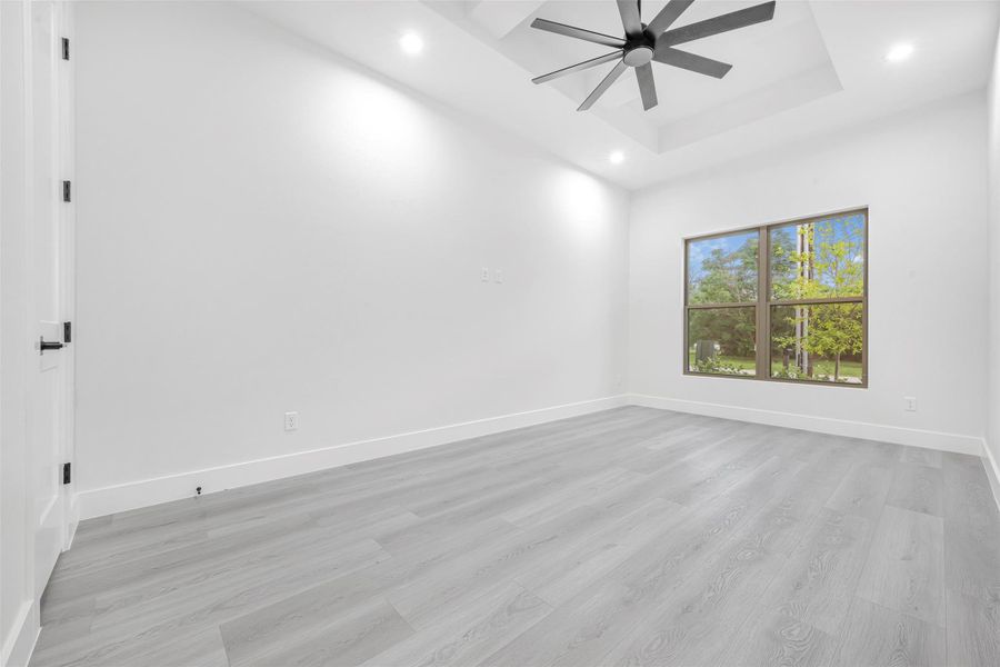 Empty room featuring a ceiling fan, recessed lighting, light wood-finished floors, raised ceiling, and baseboards. Empty room featuring a ceiling fan, recessed lighting, light wood-finished floors, raised ceiling, and baseboards.