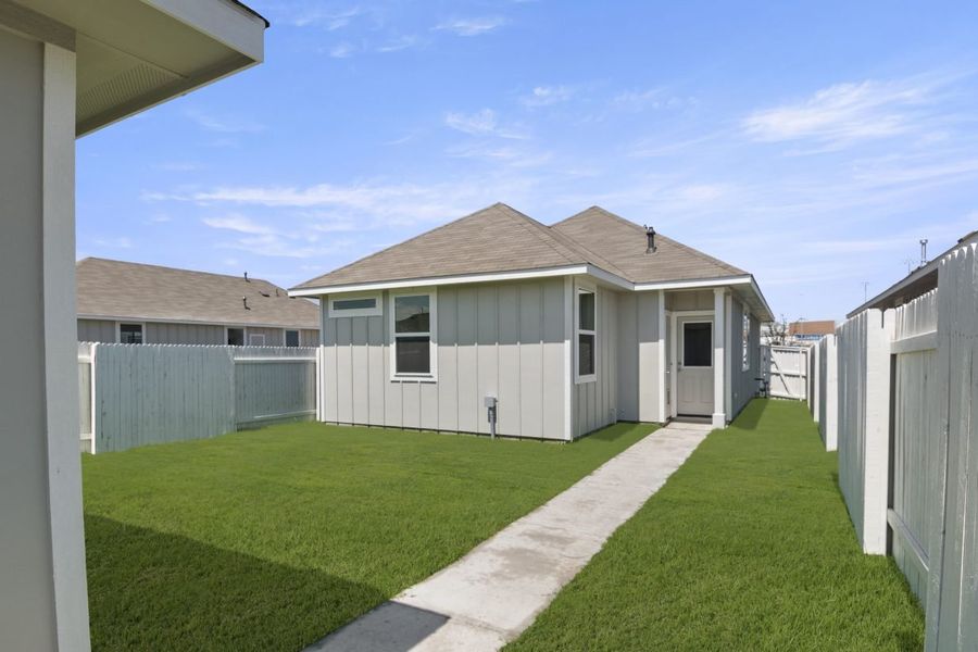 A backyard with a concrete walkway surrounded by grass leading to a gray building, and a white fence.
