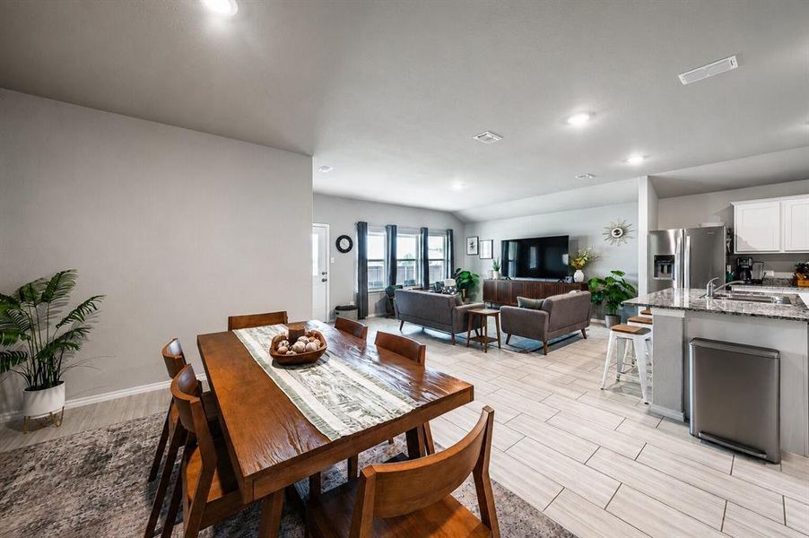 Open-concept living space featuring a dining area with a rectangular wood table, light-toned wood-finish flooring, recessed lighting, and a kitchen island with a granite-style countertop