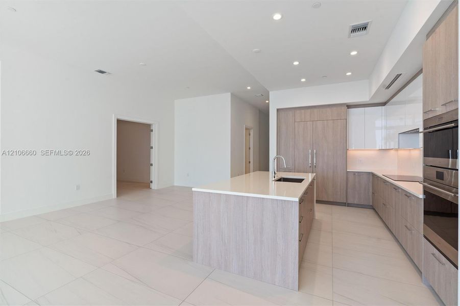 Kitchen featuring wood-finish cabinetry, white high-gloss upper cabinets, solid-surface countertops, large kitchen island with integrated sink, and built-in stainless steel double ovens
