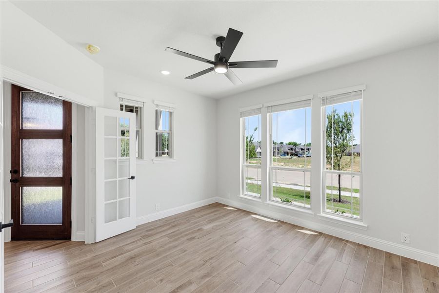 Empty room with light wood floors, ceiling fan, large windows, and dark wood entry door with glass panels Empty room with light wood floors, ceiling fan, large windows, and dark wood entry door with glass panels