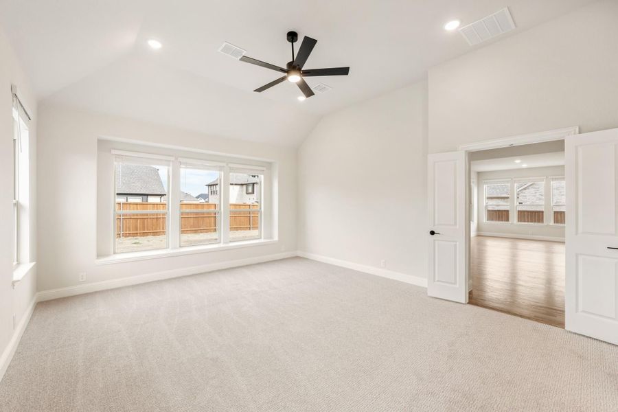 Empty bedroom with vaulted ceiling, ceiling fan, carpet flooring, and large windows overlooking backyard fence