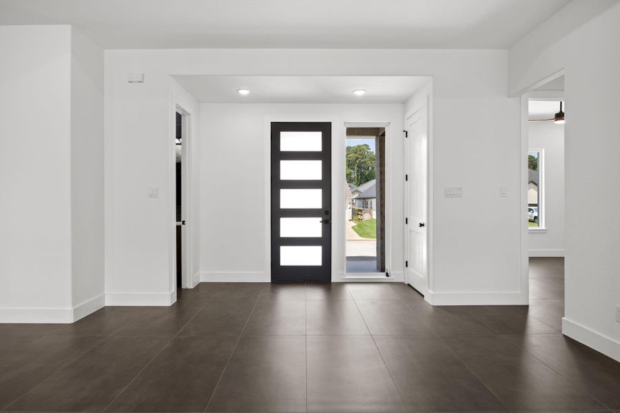 This photo shows a modern entryway with a sleek black door featuring horizontal glass panels. The space is bright and open, with large brown tiles and neutral white walls, creating a welcoming and contemporary atmosphere.