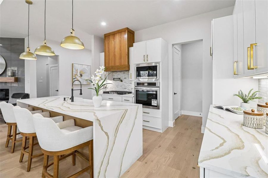 Gourmet kitchen featuring a waterfall island with striking veining, custom pendant lighting, stainless steel double ovens, a built-in microwave, and wood-finish flooring
