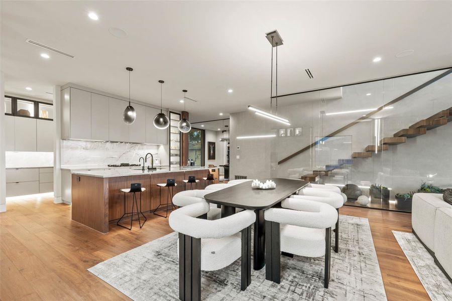 Open-concept living area featuring wood-finish flooring, a contemporary kitchen island with seating, minimalist cabinetry, marble-patterned countertops and backsplash, and a floating staircase with glass railings
